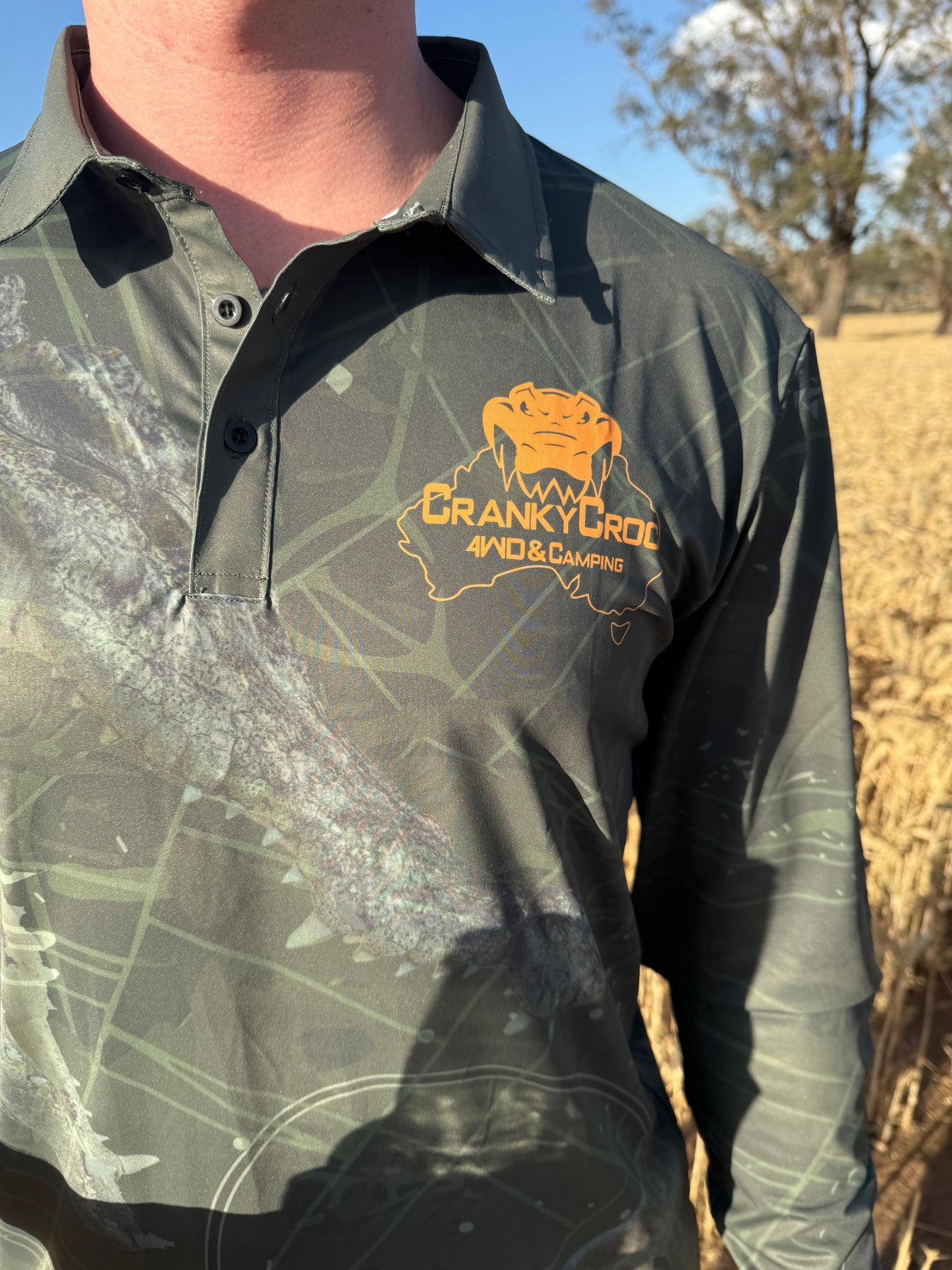 Person wearing a green camo fishing shirt / hunting shirt with an orange logo and text. The fish shirt also has a crocodile and barramundi design. The man is standing outdoors.