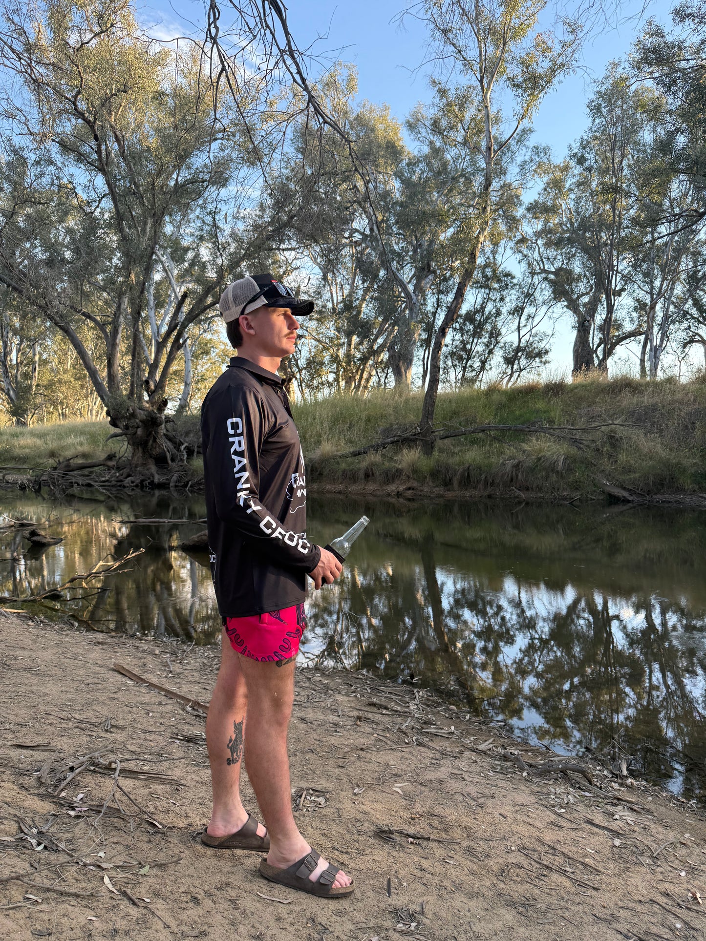 Man standing by a river holding a fishing rod, wearing a black long sleeve fishing shirt with white 'Cranky Croc' text down the sleeves and wearing pink Aussie Footy Shorts