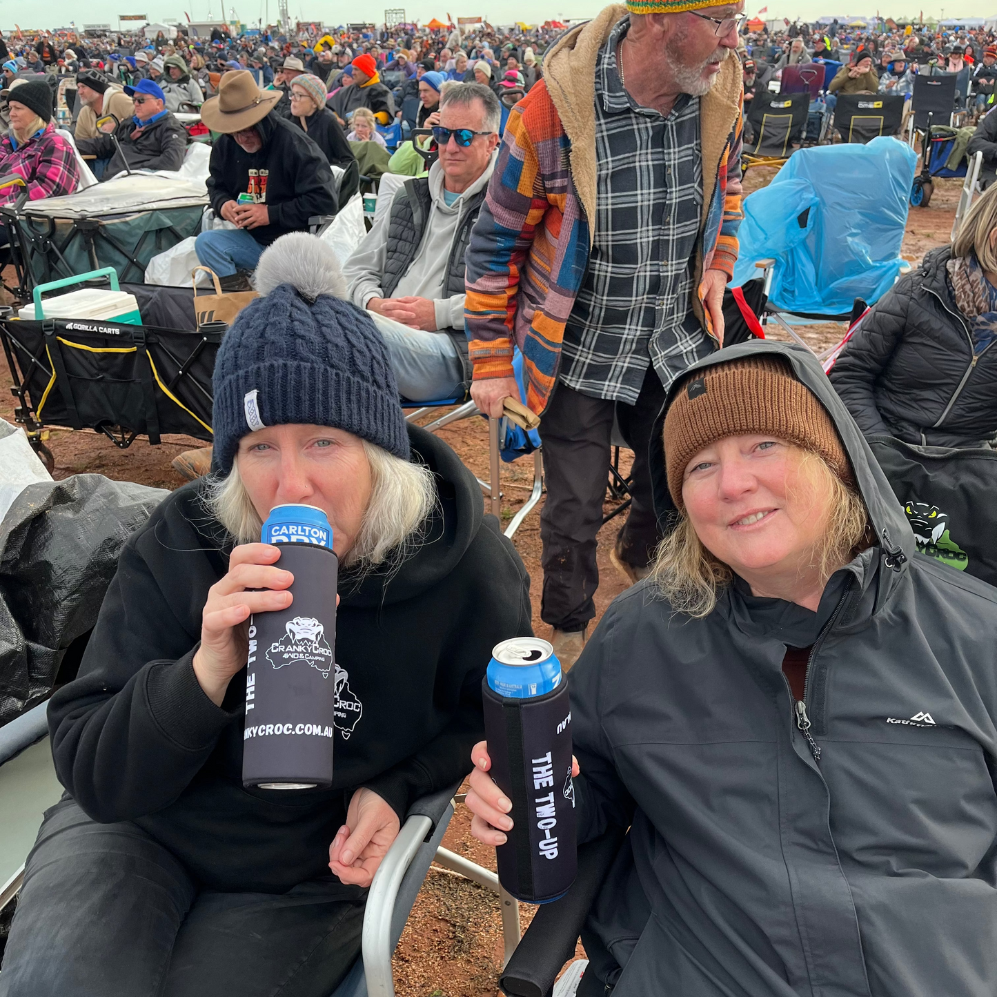 Two woman sitting on Cranky Croc camp chairs at a concert drinking beer from their two can, stubby holders