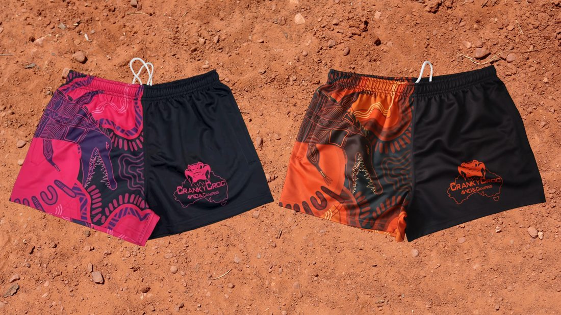 Two pairs of Indigenous Footy Shorts. One pair is orange with black the other is pink with black. The Aussie Footy Shorts with zip pockets are on a red dirt background.