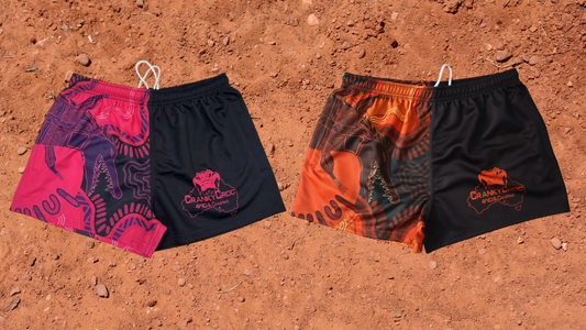 Two pairs of Indigenous Footy Shorts. One pair is orange with black the other is pink with black. The Aussie Footy Shorts with zip pockets are on a red dirt background.