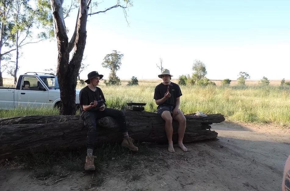 Two young men sitting on a log on farm land wearing Folding Pouch Hat