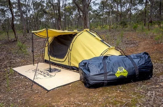 Swag sitting in bushland with swag awning set up.  In front of the swags a black PVC swag bag. Next to the swag under the awning is a swag mat.