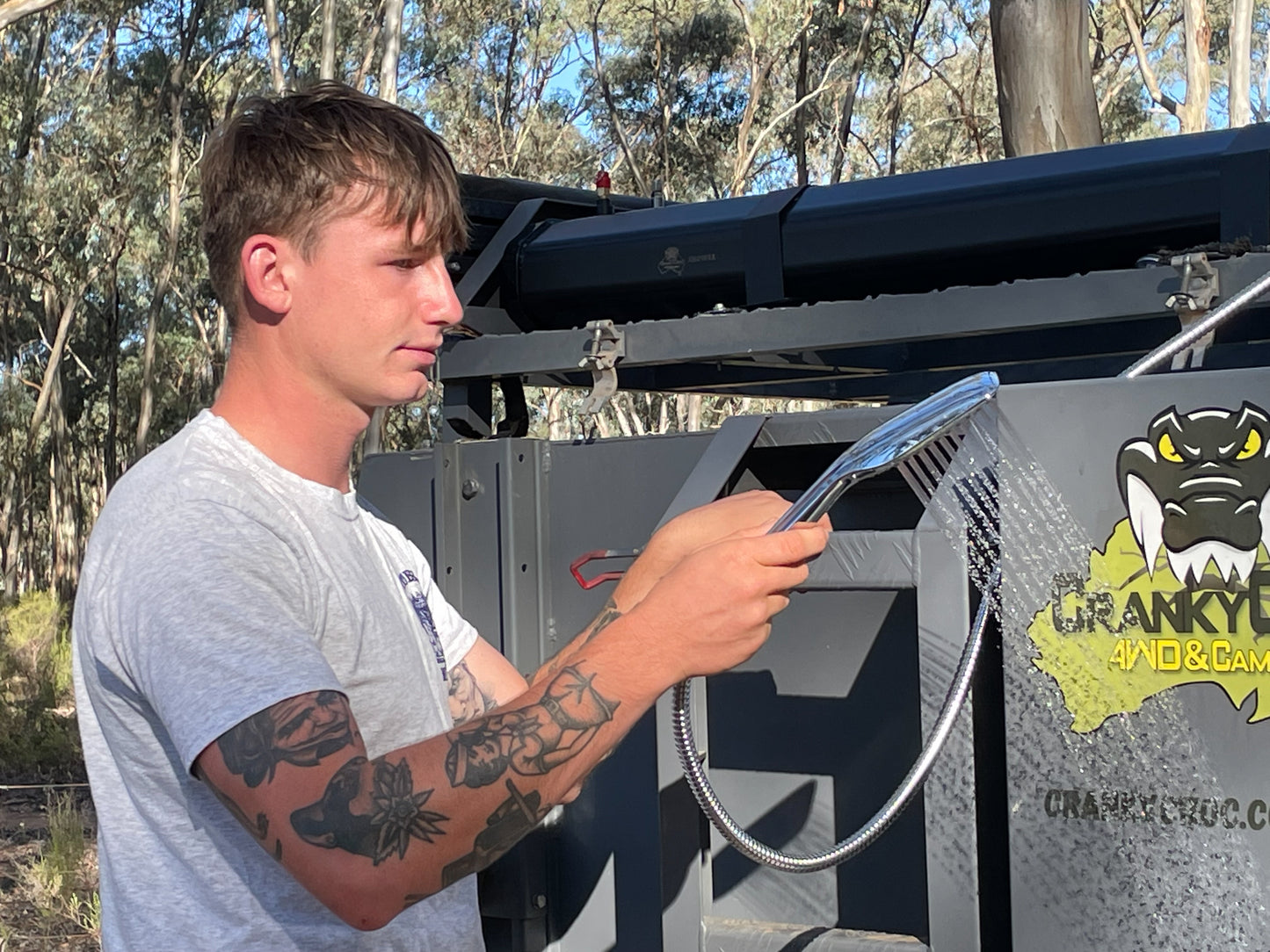 Young man using shower head from roof mounted solar shower system in bush land.