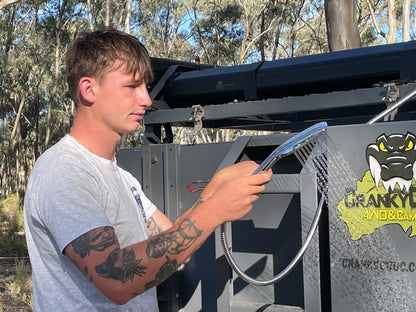 Young man using shower head from roof mounted solar shower system in bush land.
