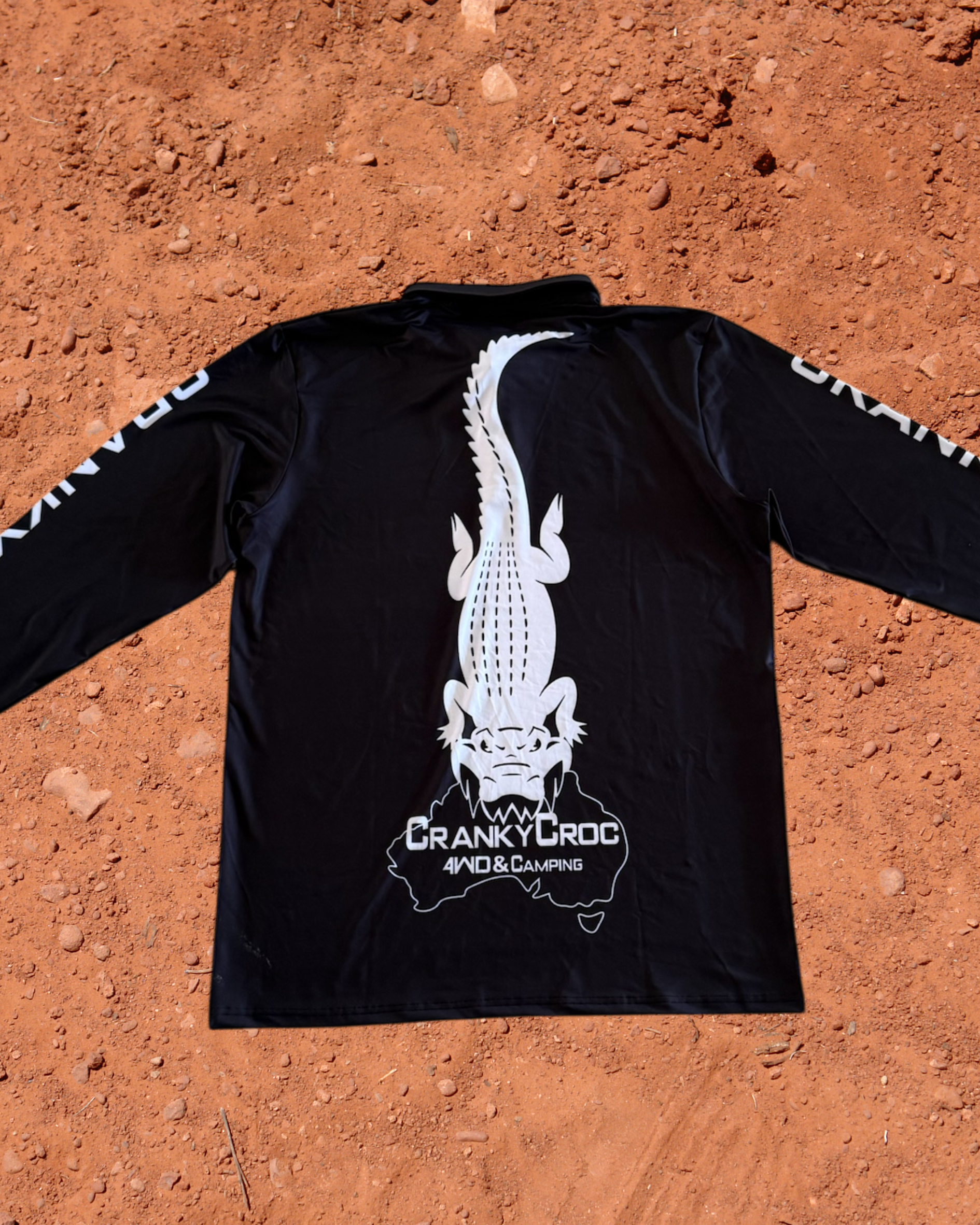 Back of a black long sleeve fishing shirt, the fish shirt has white text on the sleeve with a white Cranky Croc logo on the back. The logo is a crocodile biting down on Australia. Shirt is on a red dirt background
