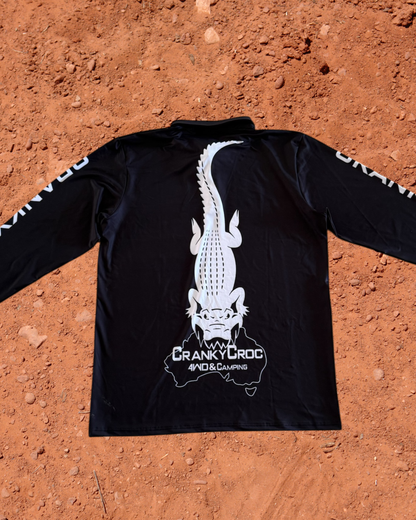 Back of a black long sleeve fishing shirt, the fish shirt has white text on the sleeve with a white Cranky Croc logo on the back. The logo is a crocodile biting down on Australia. Shirt is on a red dirt background