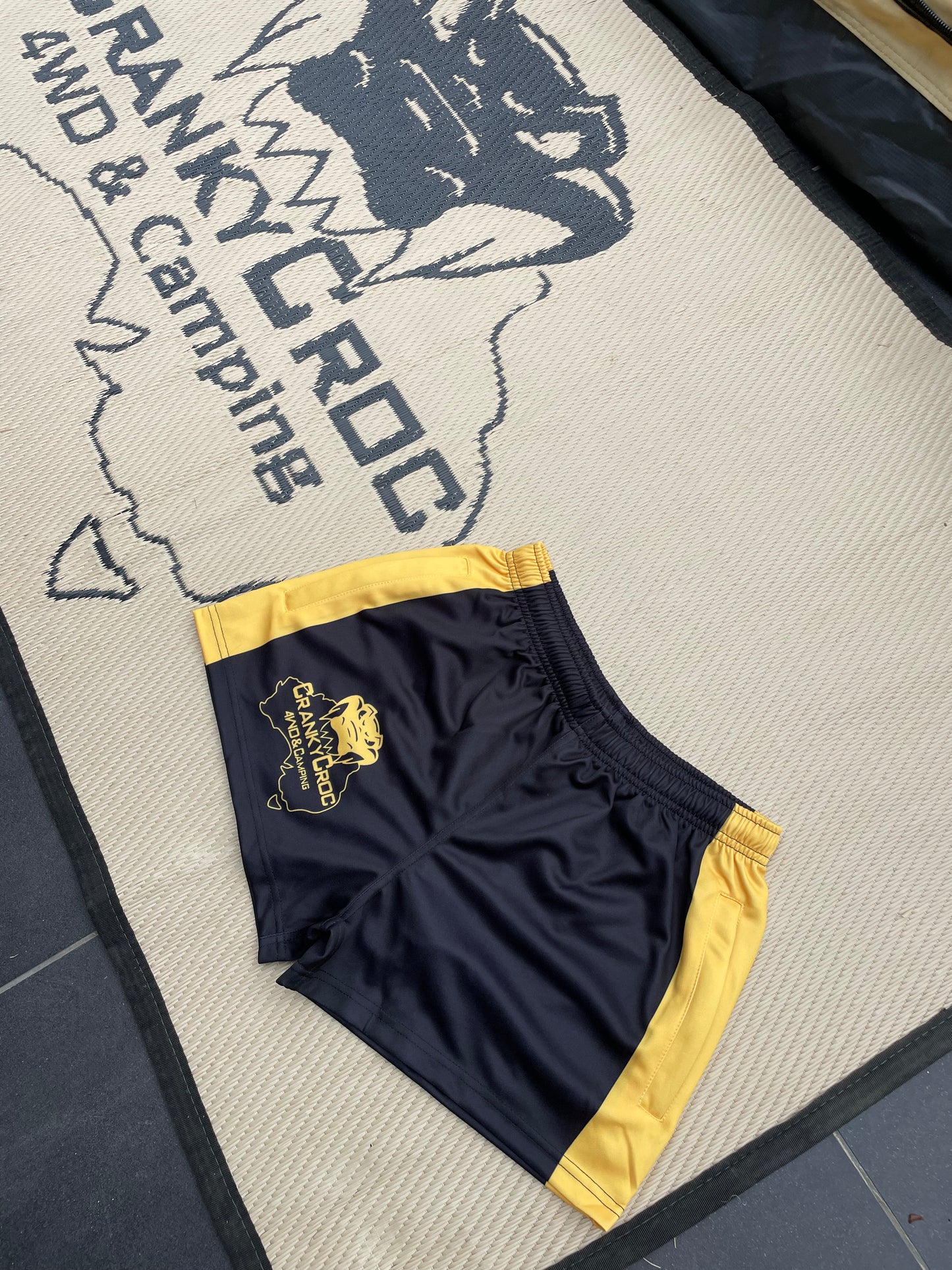Black Footy Shorts with yellow trim on a mat with 'Cranky Croc' branding.