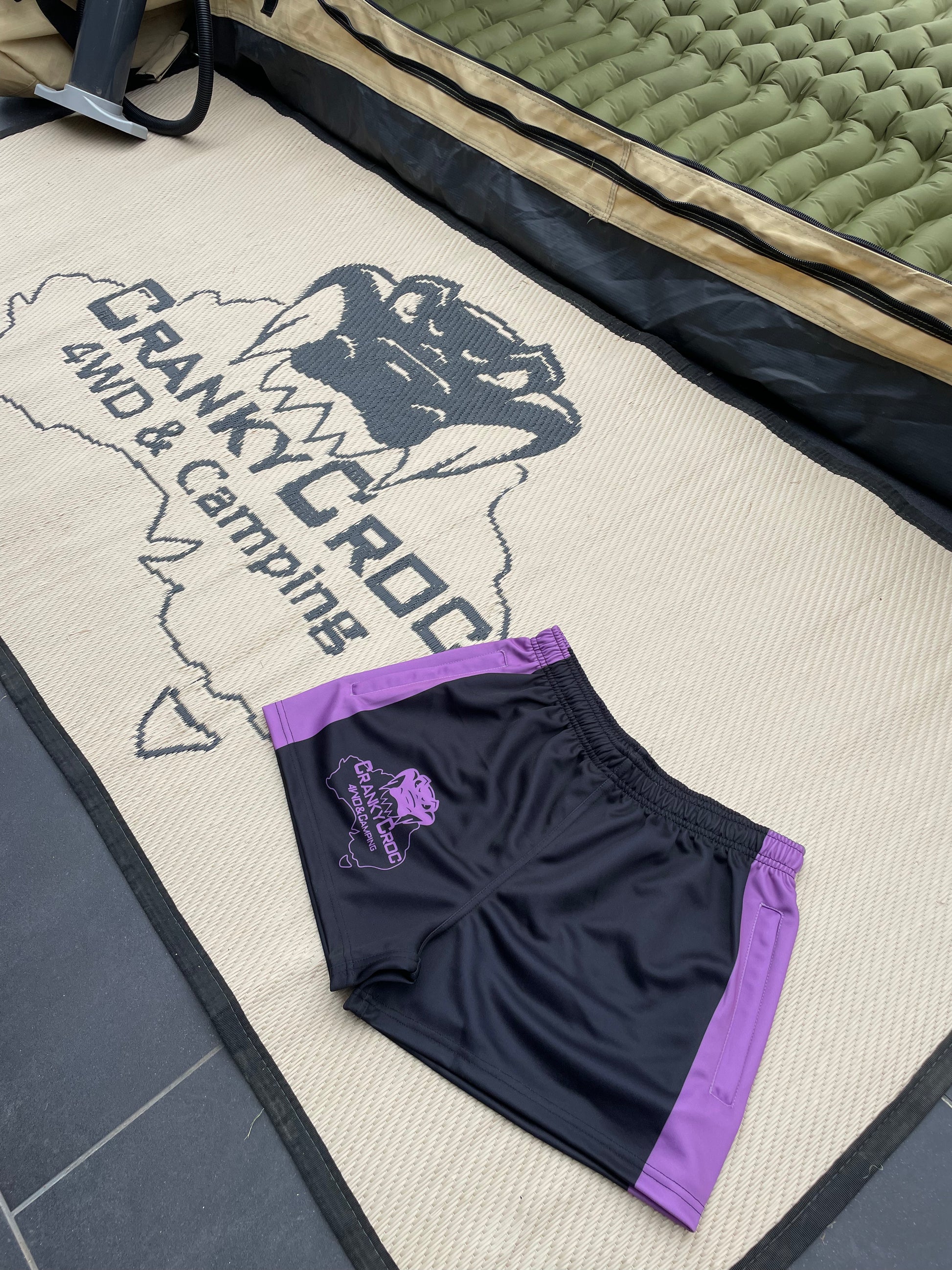 Black and purple Footy Shorts with a logo on a mat with 'Cranky Croc 4WD & Camping' branding.