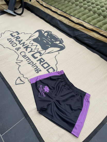 Black and purple Footy Shorts with a logo on a mat with 'Cranky Croc 4WD & Camping' branding.