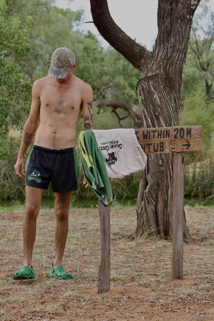 Man wearing black with green Aussie Footy Shorts with his shirt off in Aussie bushland