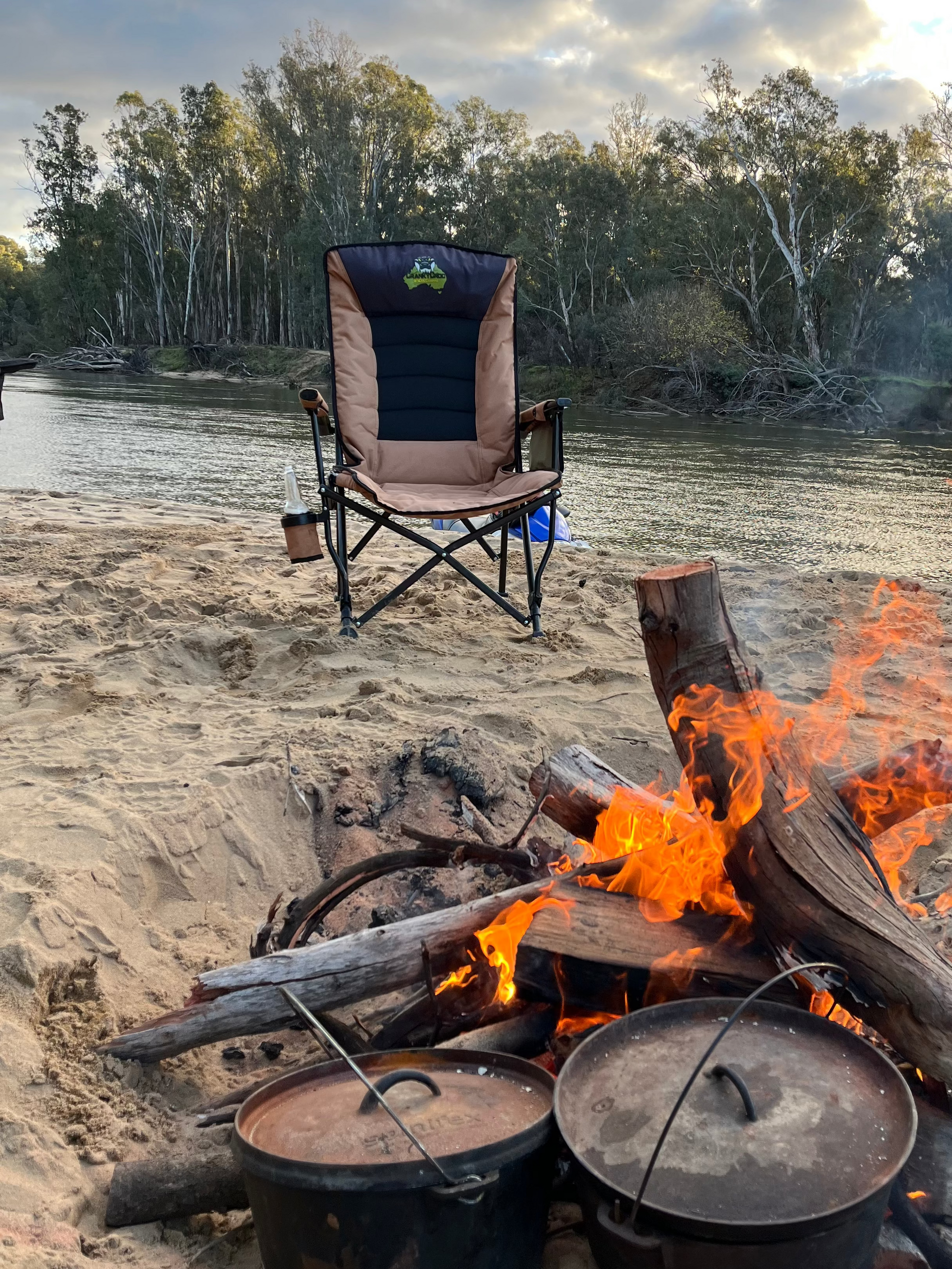 A camping chair sits on a sandy riverbank with a campfire burning in the foreground and two cast iron pots cooking over the fire. Trees line the river in the background under a cloudy sky.