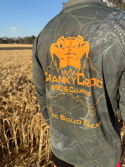 Person wearing a green camo fishing shirt / hunting shirt with orange branding in a field. The fish shirt has a crocodile and barramundi fish design.