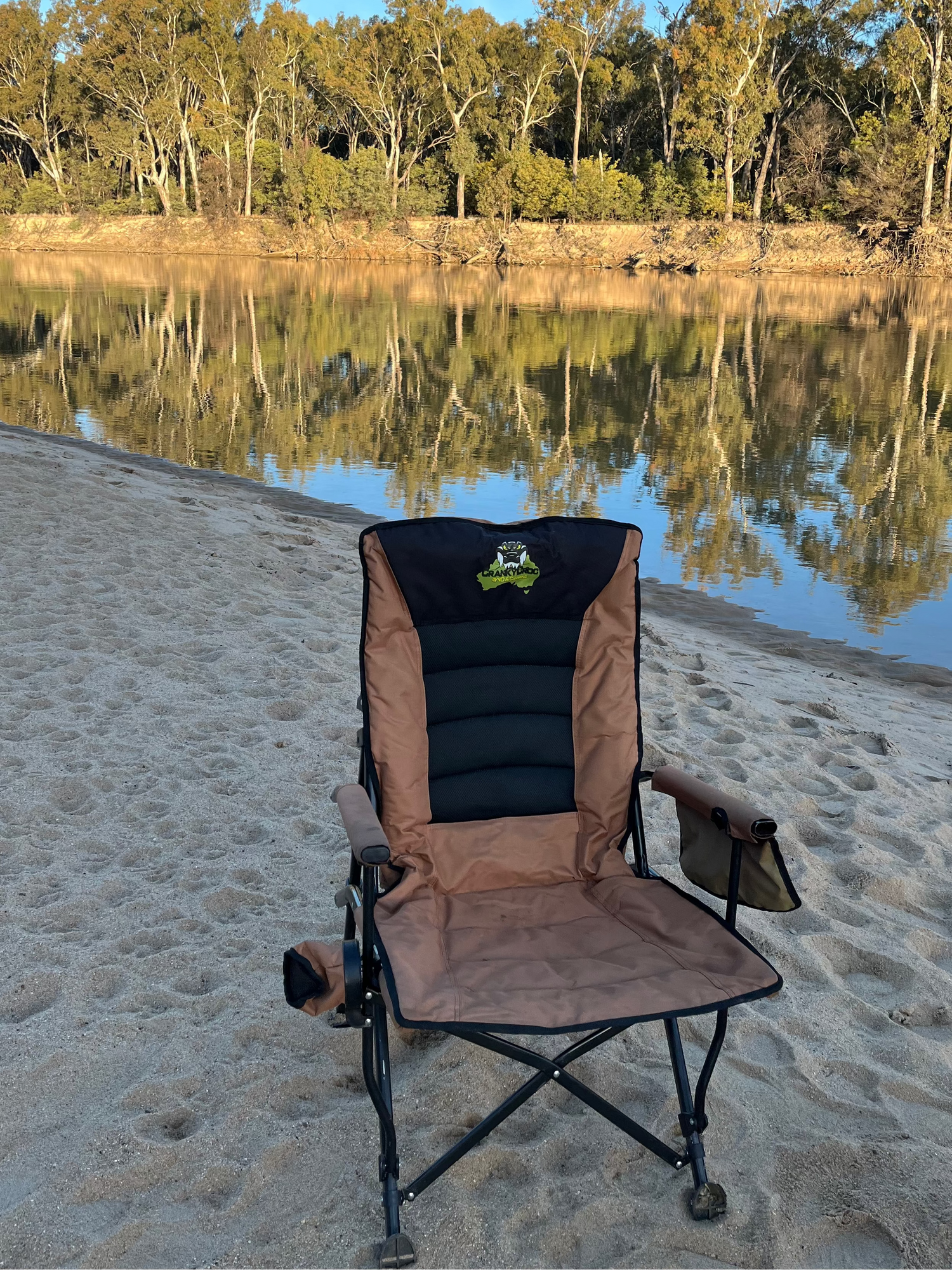 Folding chair on a sandy beach with a calm river and trees in the background