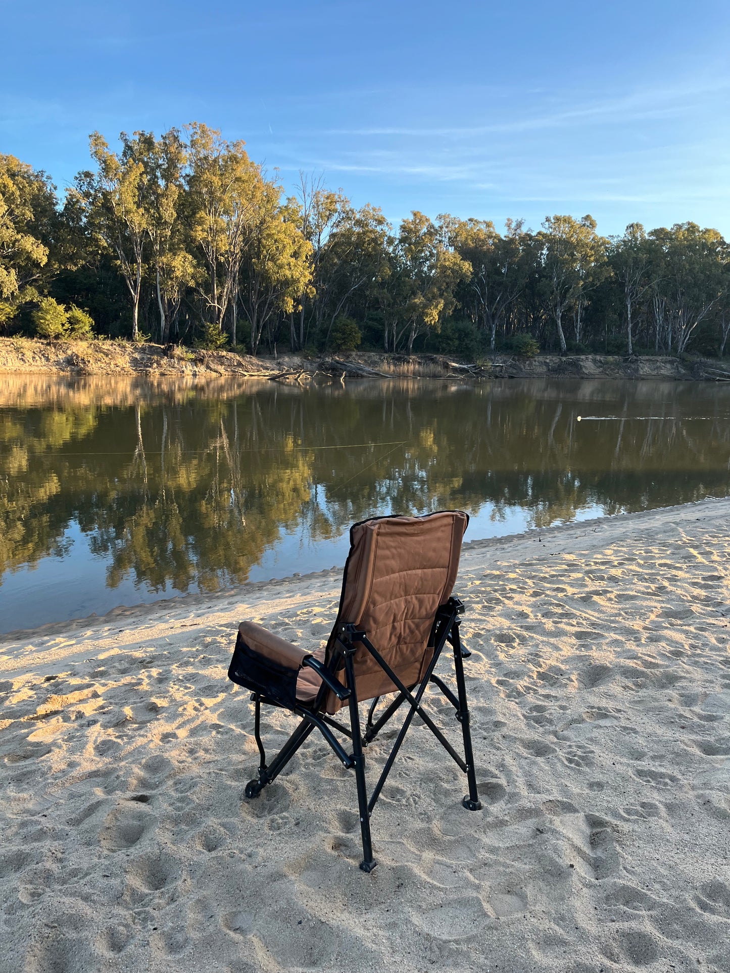 Brown camping chair on a sandy beach with a body of water and trees in the background