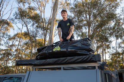 A man standing on a roof rack next to a black PVC swag bag, with trees in the background.