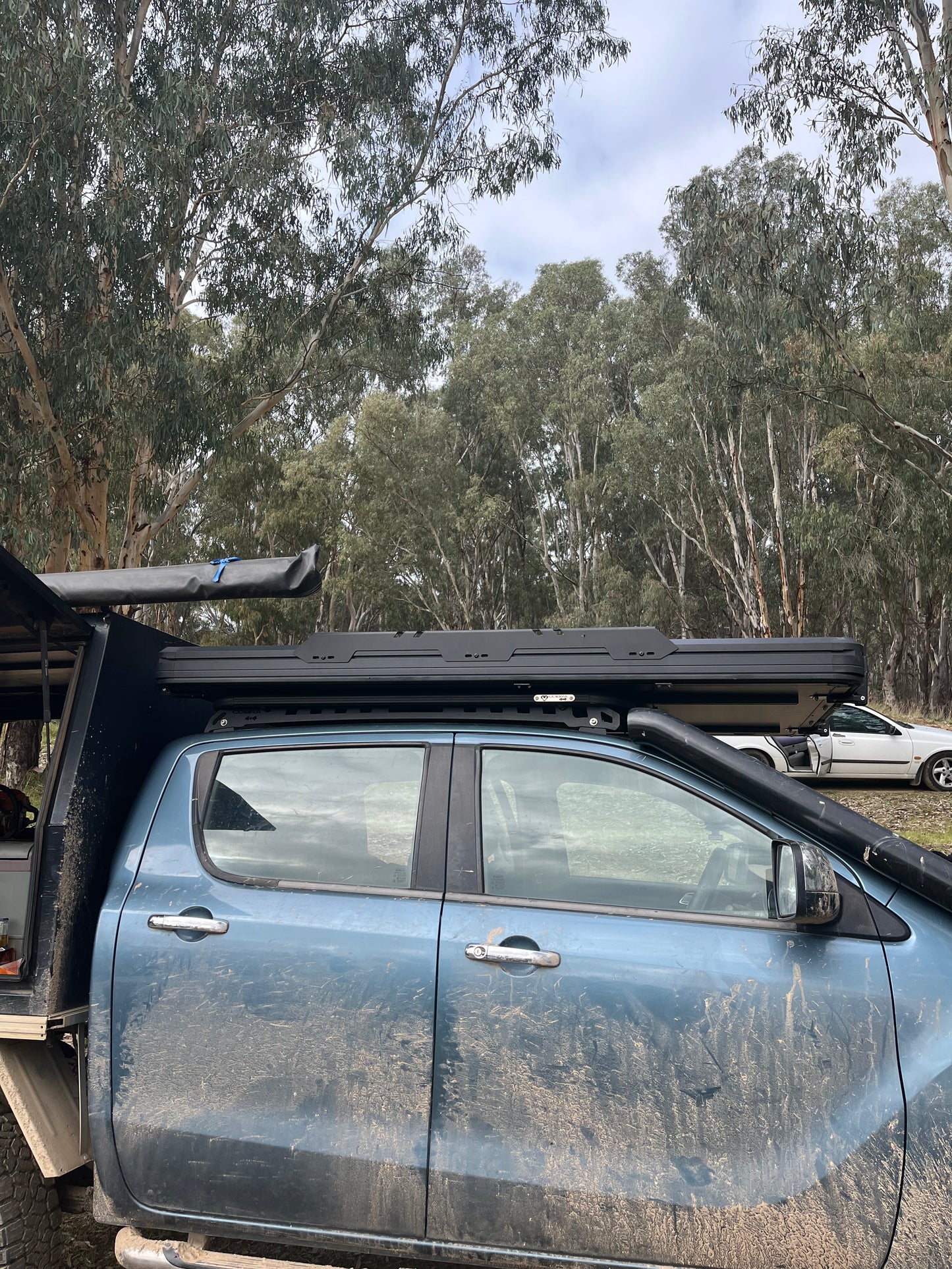 A dusty blue 4WD truck with a black hardtop rooftop tent and roof rack, parked outdoors in a wooded area with trees in the background.