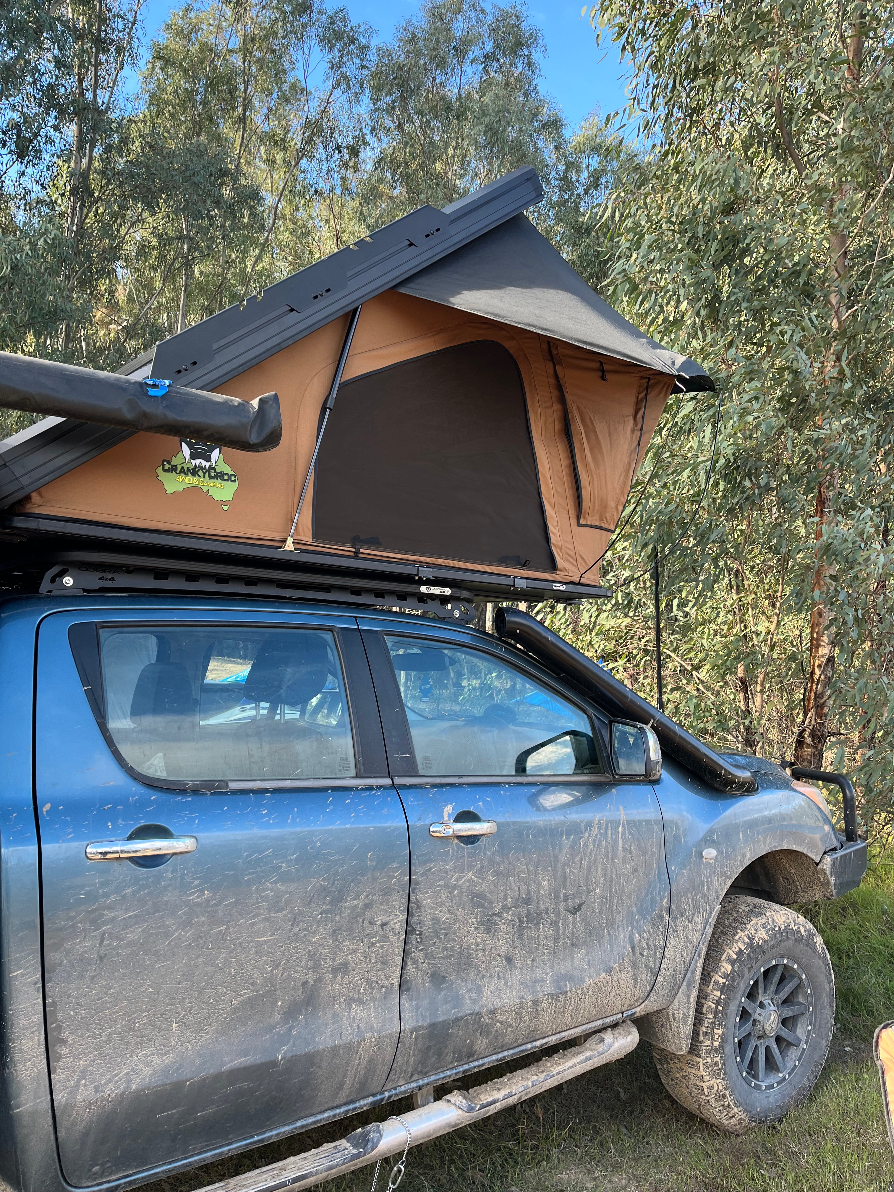 A Cranky Croc rooftop tent in tan and black mounted on the roof rack of a dusty blue 4WD truck, parked outdoors in a wooded area with trees and blue sky visible.