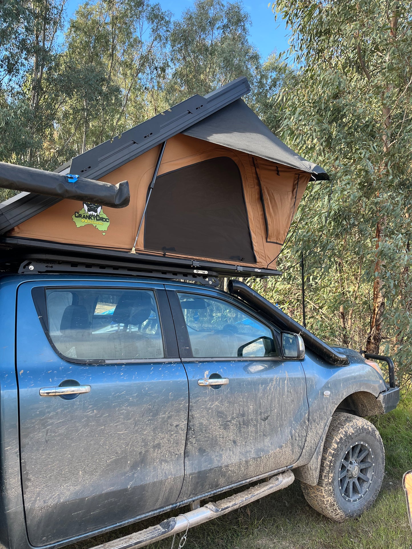 A Cranky Croc rooftop tent in tan and black mounted on the roof rack of a dusty blue 4WD truck, parked outdoors in a wooded area with trees and blue sky visible.