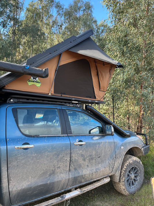 A Cranky Croc rooftop tent in tan and black mounted on the roof rack of a dusty blue 4WD truck, parked outdoors in a wooded area with trees and blue sky visible.
