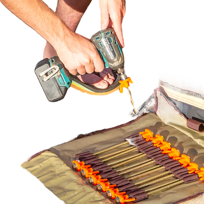 A person is using a hand drill to drive 'screw in tent pegs' into the ground, with the beige roll-up bag of pegs visible in the foreground.