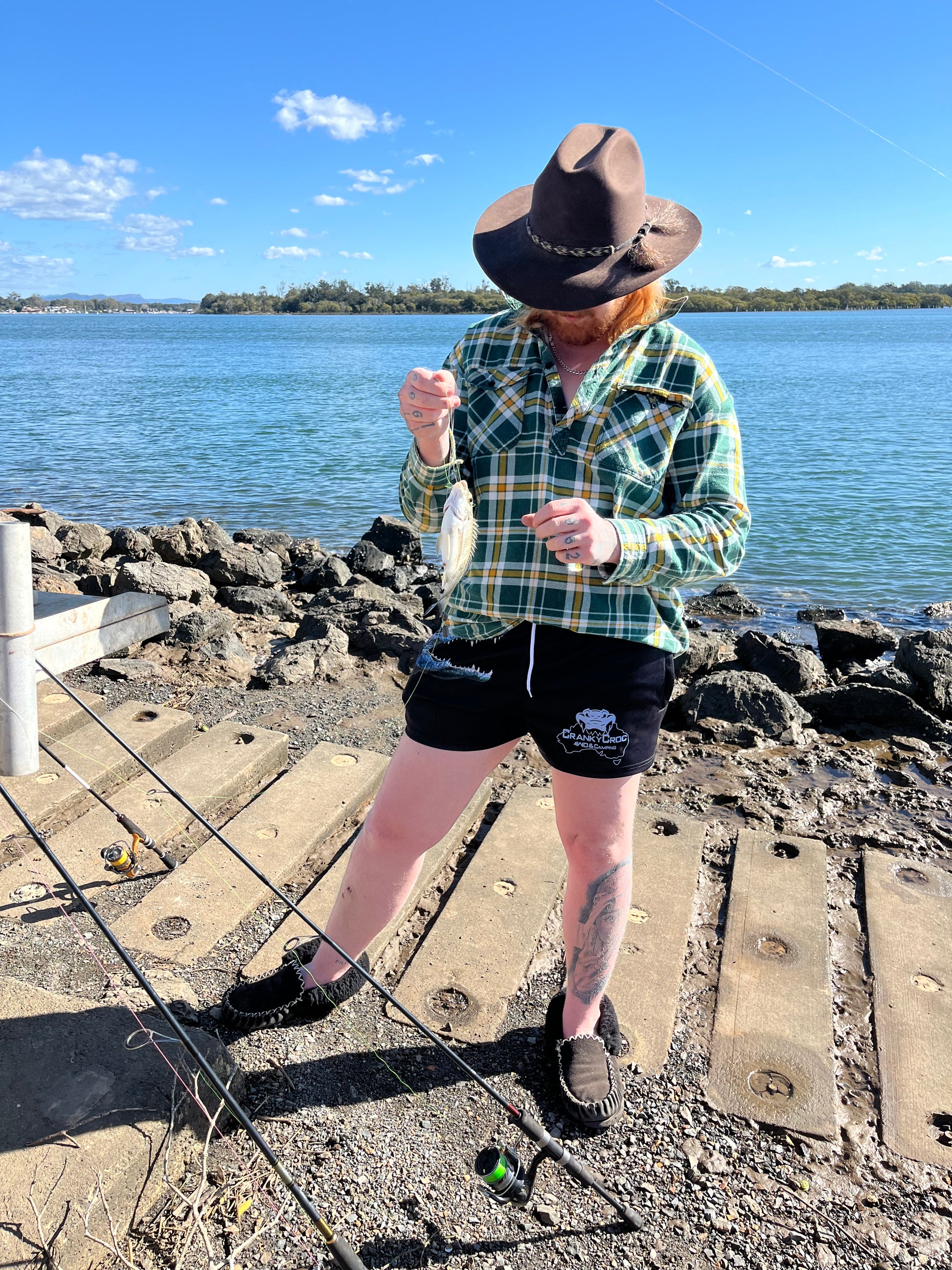 Person fishing by a body of water wearing a hat and wearing Aussie Footy Shorts