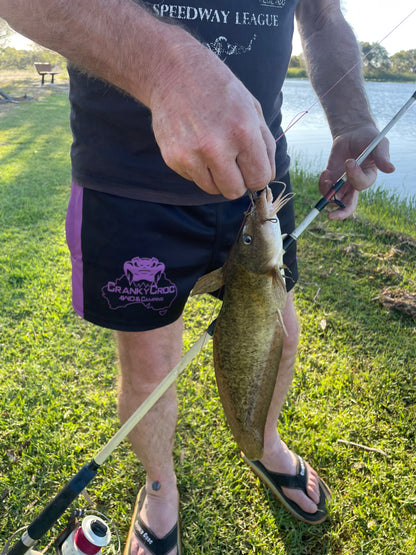 Person holding a fish caught on a fishing rod by a lake while wearing black with purple Aussie Footy Shorts