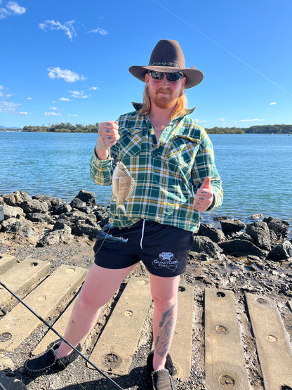 Person holding a fish by a body of water on a sunny day wearing black Aussie Footy Shorts with a crocodile on the front