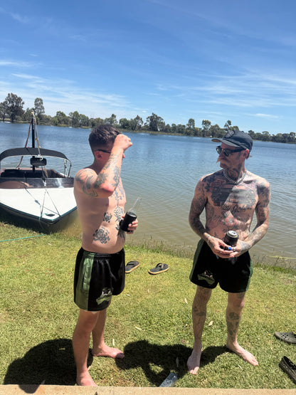 Two men with tattoos standing by a lake with boats in the background wearing green with black Aussie Footy Shorts