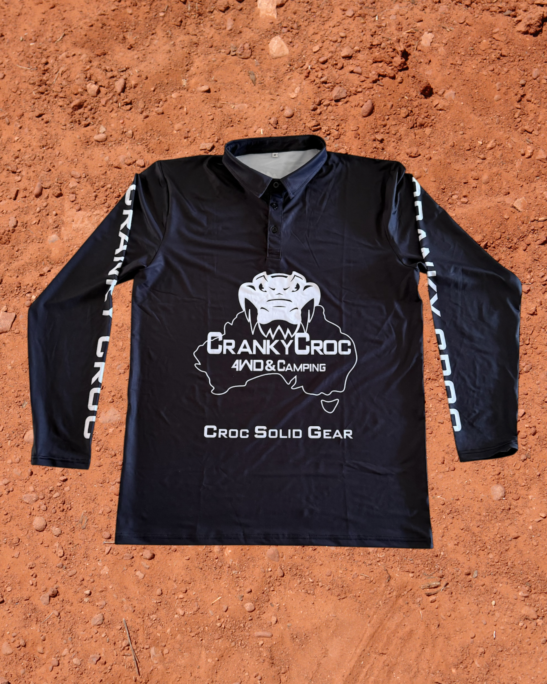 Front of black long sleeve fishing shirt, the fish shirt has white text on the sleeve with white Cranky Croc branding on the front. The logo is a crocodile biting down on Australia. Shirt is on a red dirt background