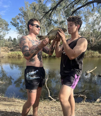 Two people holding a fish by a river with trees in the background wearing Cranky Croc Aussie Footy-Shorts