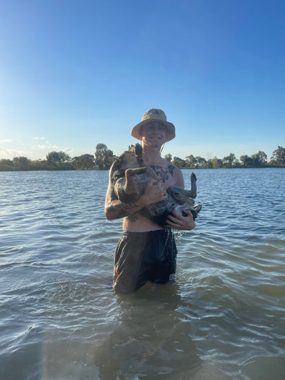 Young man standing in lake wearing a Folding Fishing Pouch Hat holding blue heeler pup.