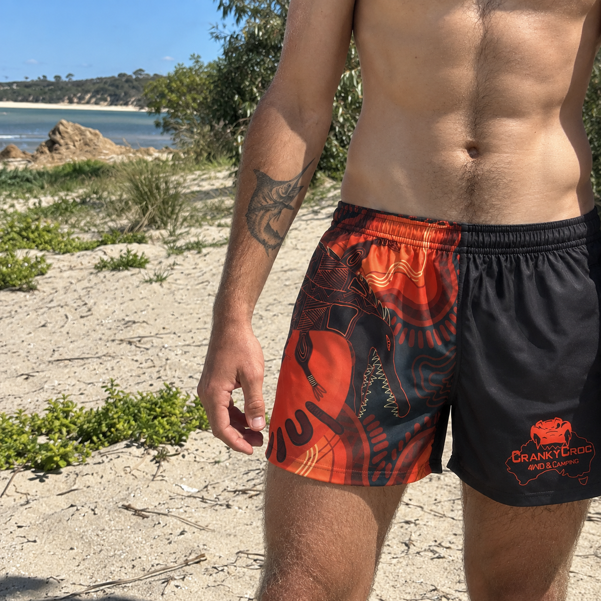 A man wearing Aussie indigenous footy shorts while standing on a sandy beach with ocean and trees in the background.  