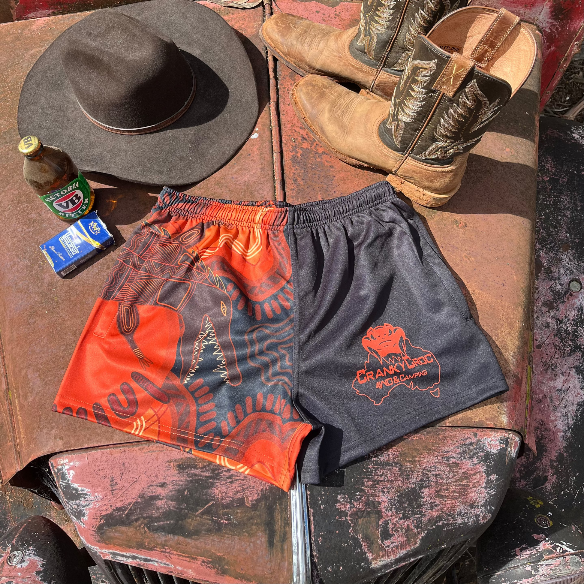 Indigenous Country Aussie Footy Shorts on a 4wd bonnet next to a pair of country boots, country cowboy hat and a beer.