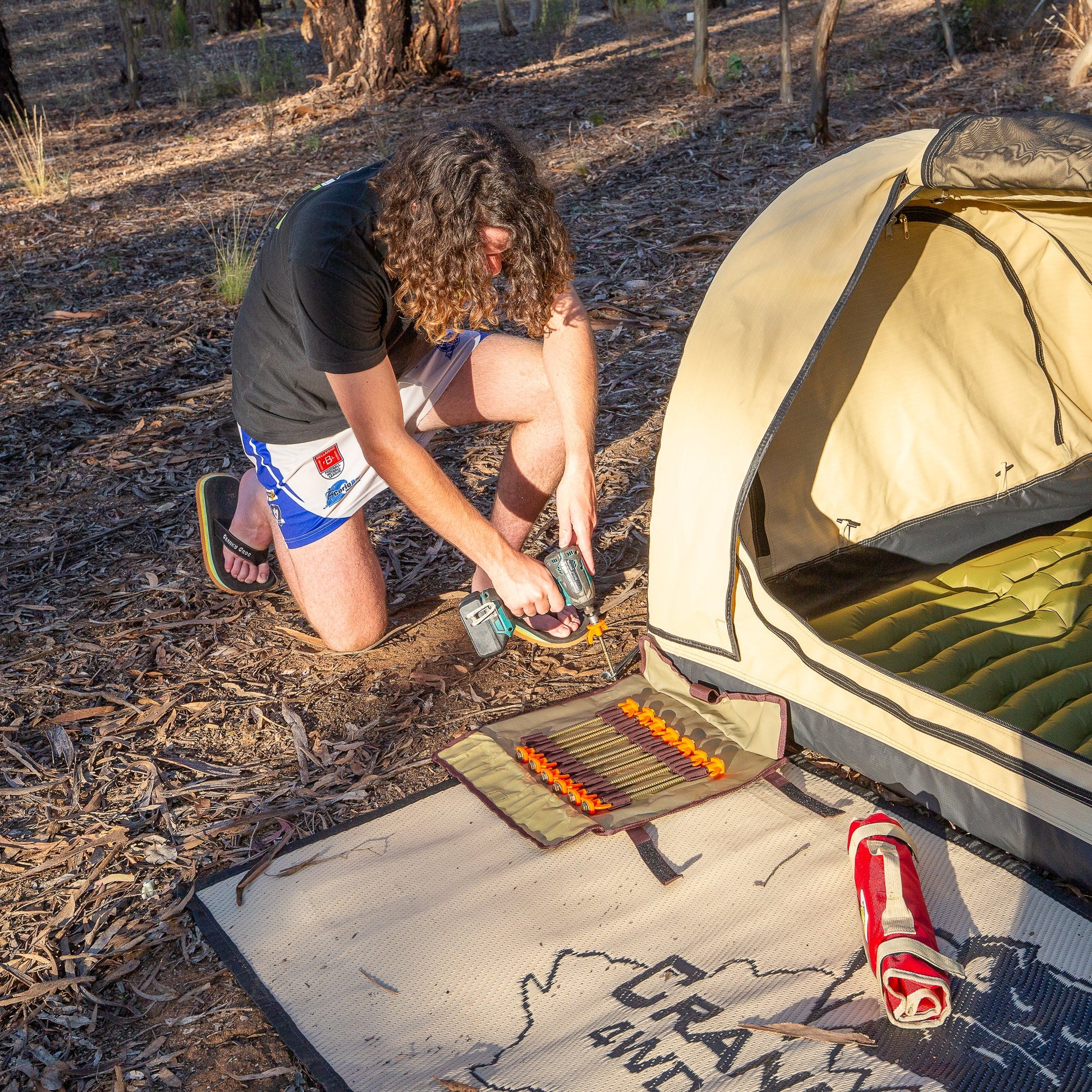 Man boy crouching next to Inflatable swag drilling 'screw in tent pegs' into ground for inflatible swag 