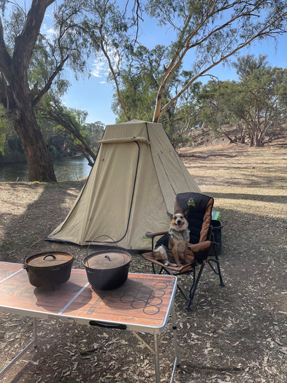 Camping scene with a family tent, table, and dog sitting on a camp chair. The instant tent is set up in bushland by a river
