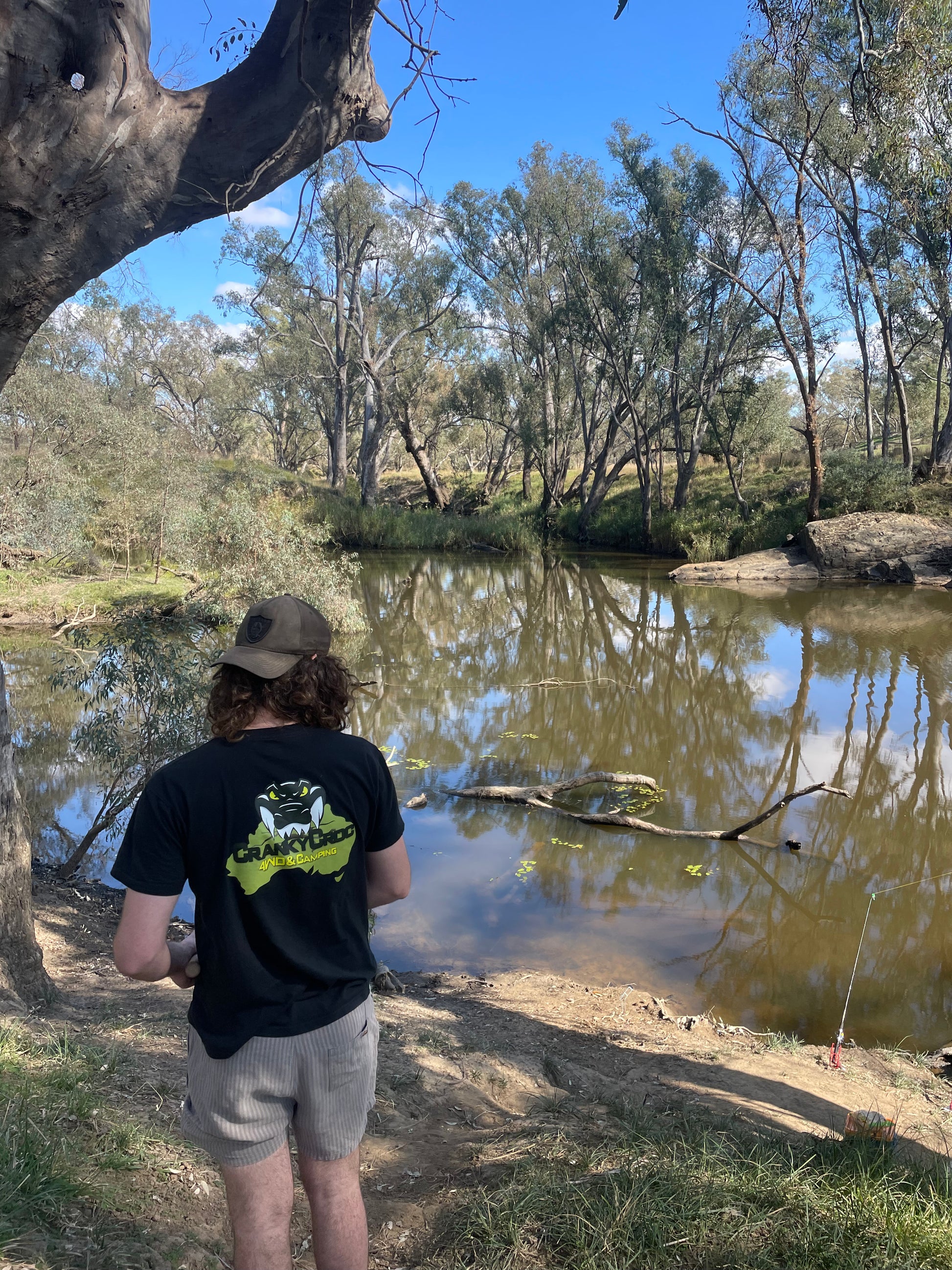 Person fishing by a river with trees and clear sky in the background wearing a black tee with a big Cranky Croc logo on the back