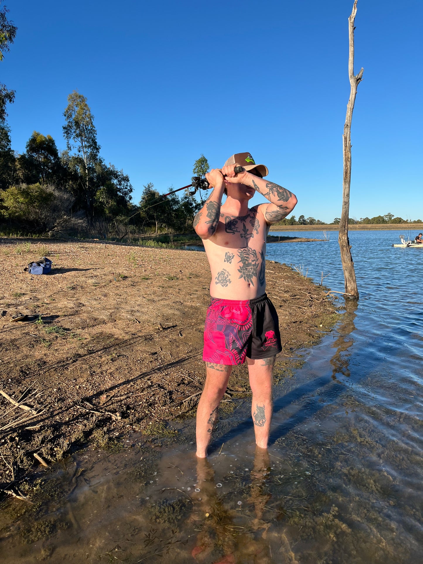 Man standing in shallow water by a lake, holding a fishing rod while wearin black with pink indigenous footy shorts with zip pockets and a crocodile design
