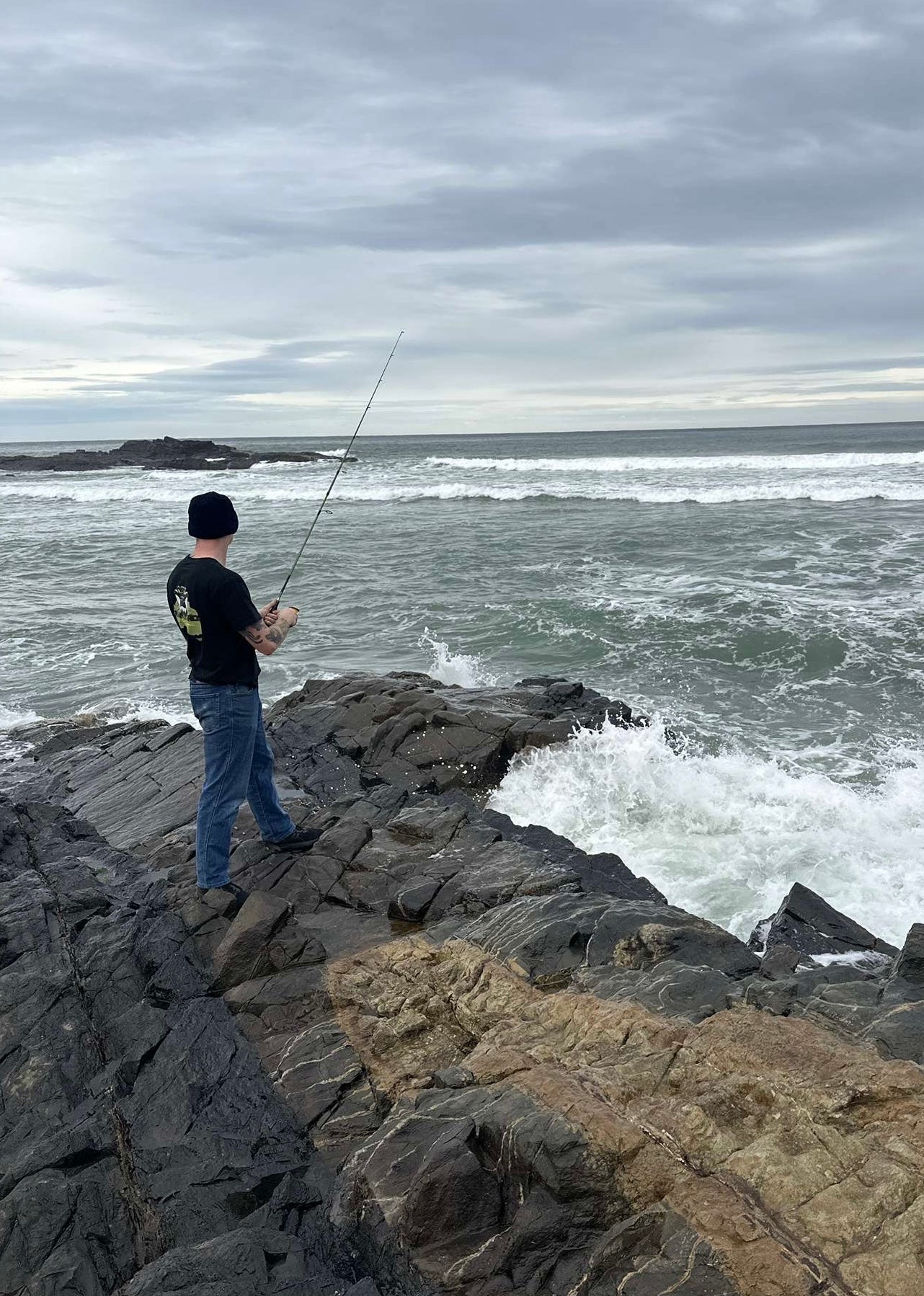 Person fishing on rocky coastline with ocean waves and cloudy sky wearing a black tee branded Cranky Croc 4WD and Camping.