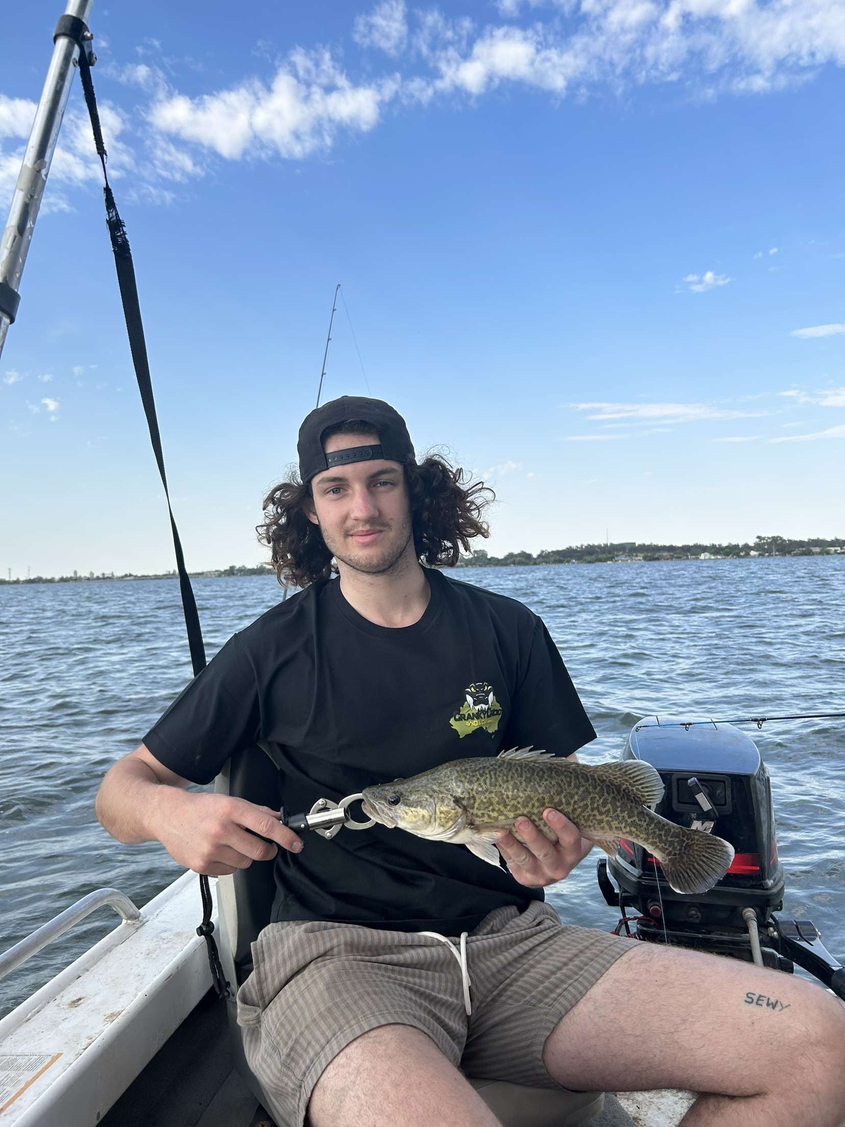 Man holding a murray cod fish on a boat with a clear blue sky and water background. the man is wearing a black tee by Cranky Croc