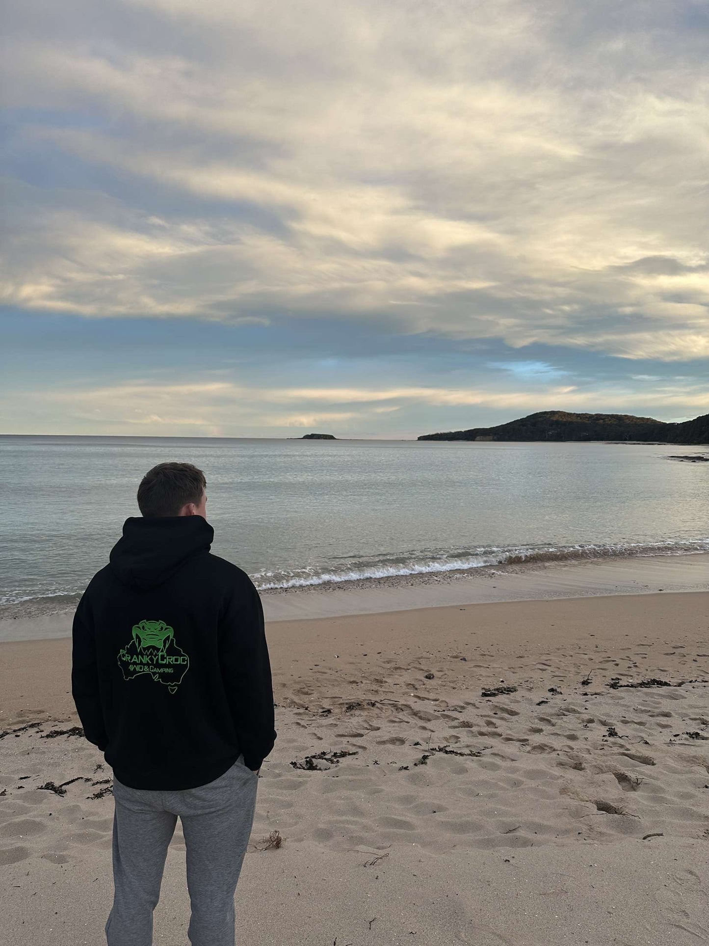 Person wearing a black hoodie by Cranky Croc, standing on a beach looking at the ocean with a cloudy sky.