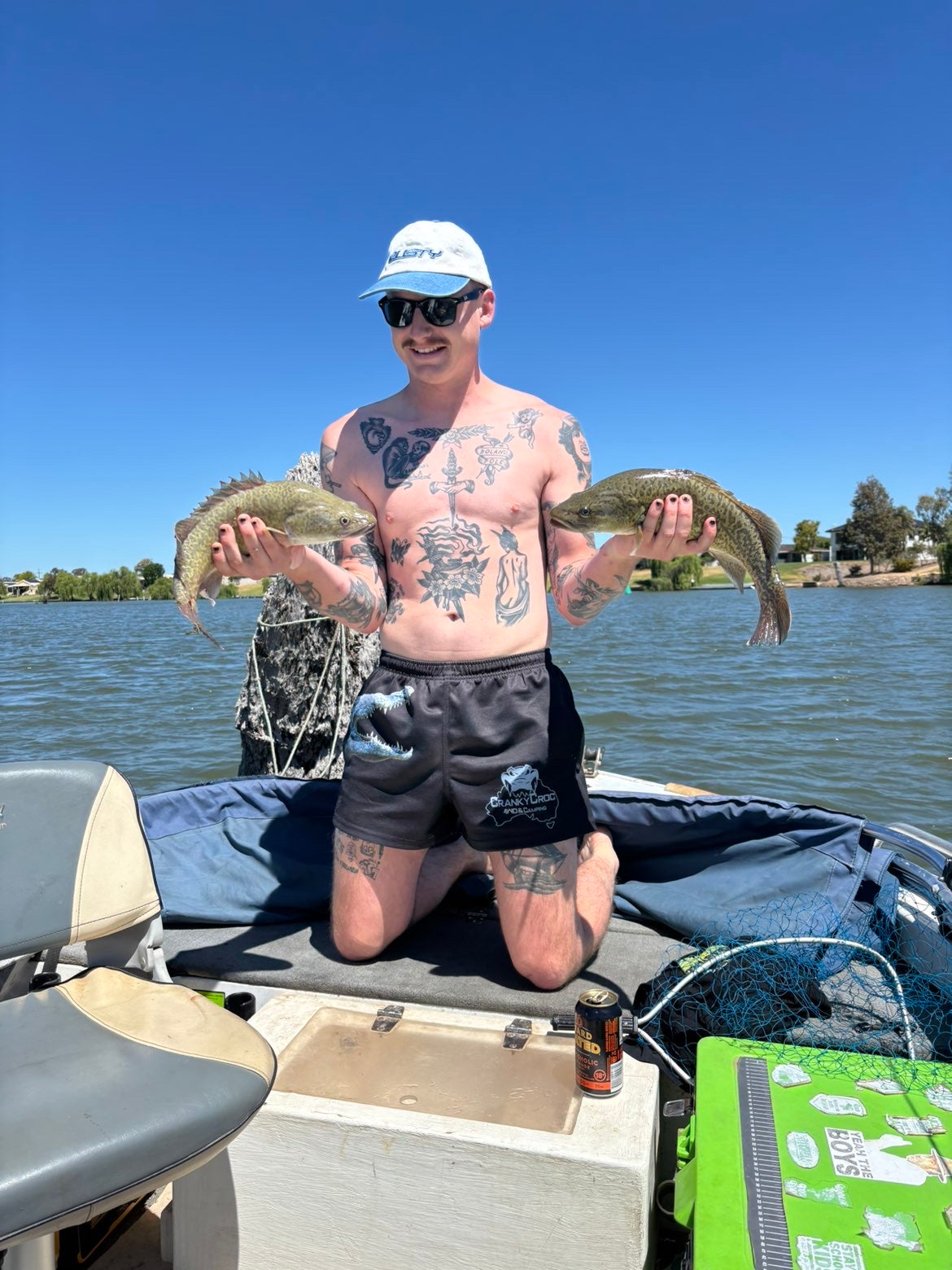 Man on a boat holding two murray cod fish with a clear blue sky wearing a pair of black Aussie Footy Shorts with a grey crocodile on the front. Footy Shorts with pockets 