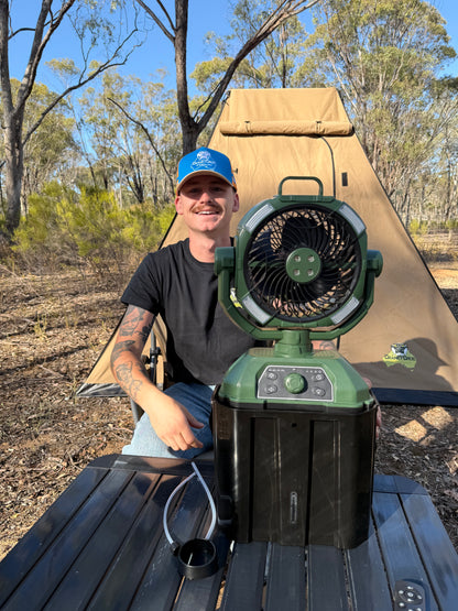 Man sitting outdoors with a large misting fan on a table, surrounded by trees and camping equipment. The camping fan is coloured green