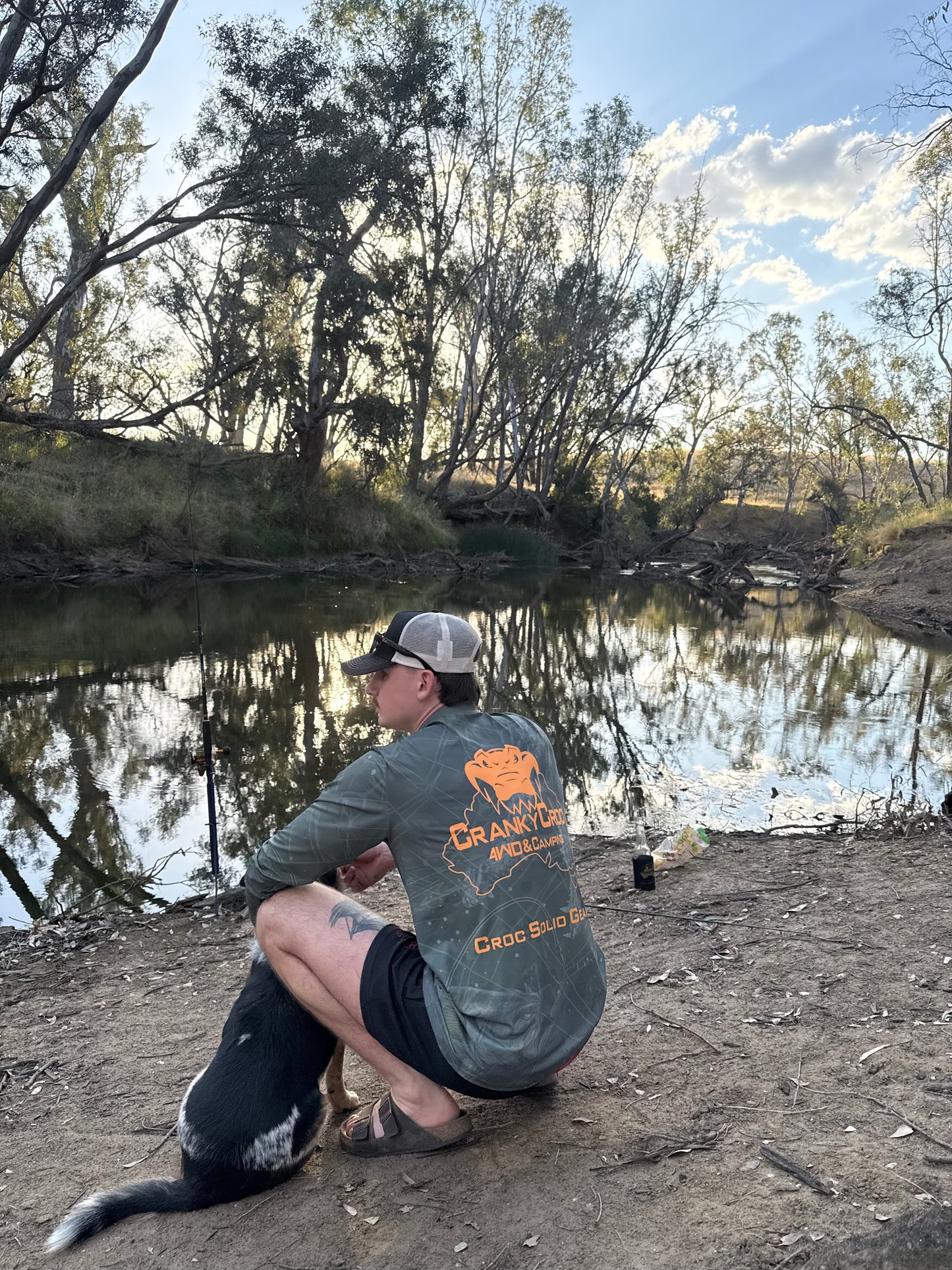 Man sitting by a river bank with a dog, wearing a branded long sleeve fishing shirt / hunting shirt with fluro orange logo. The fish shirt has a crocodile and barramundi design.