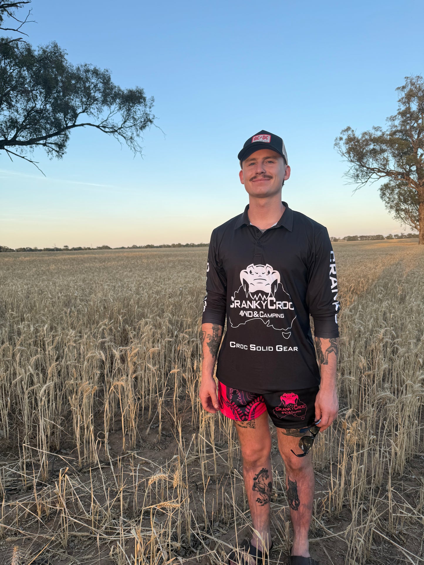 Man standing in a field wearing a black fishing shirt with crocodile design branding. The sleeves also have white text saying Cranky Croc. The man is standing in a field.