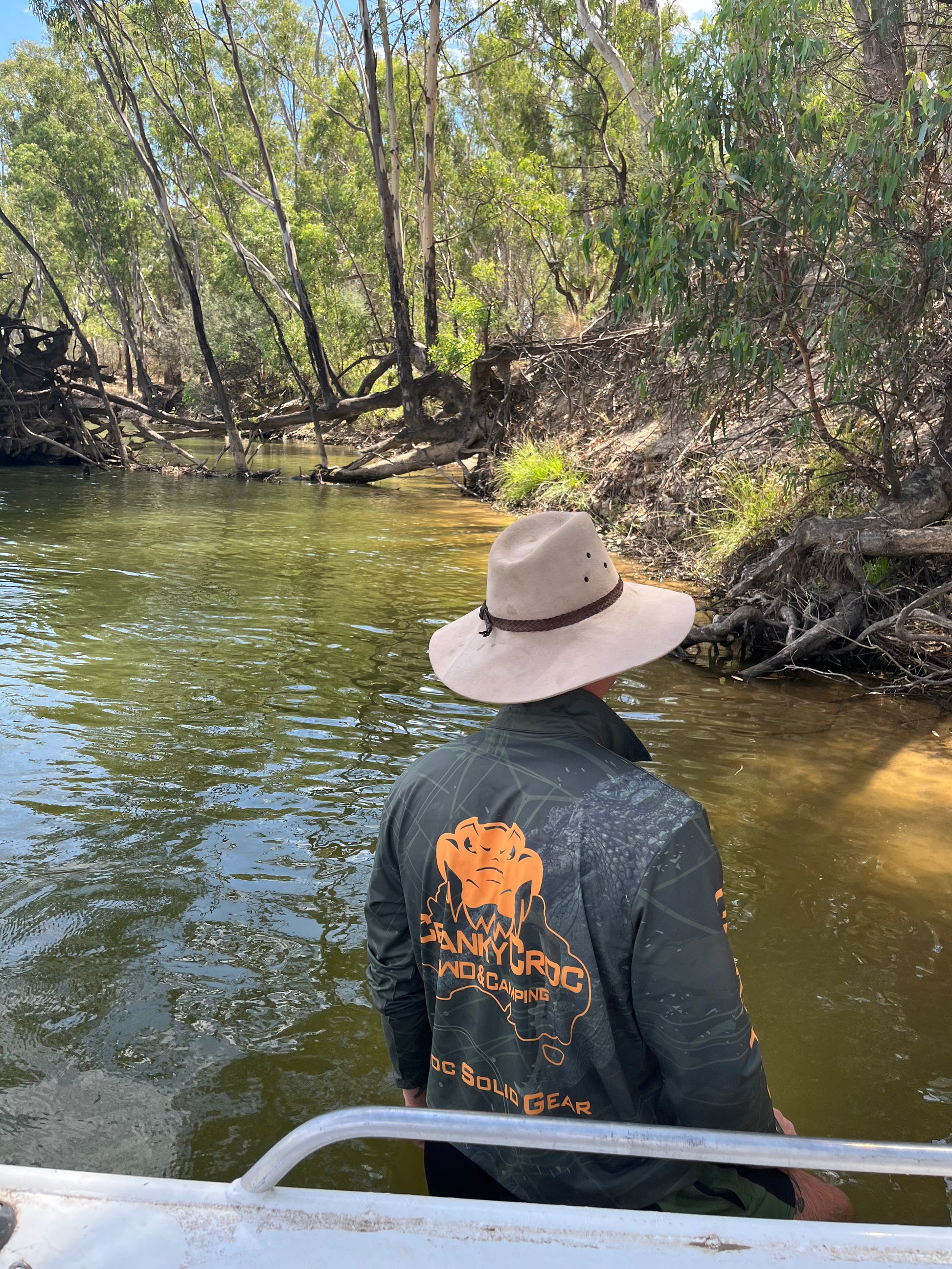Man wearing dark green long sleeve fishing shirt / hunting shirt. The fish shirt has a fluro orange 'Cranky Croc' logo on the back with a crocodile and barramundi design. Man wearing the camo fishing shirt is standing in a river next to a fishing boat and is wearing a Akubra hat