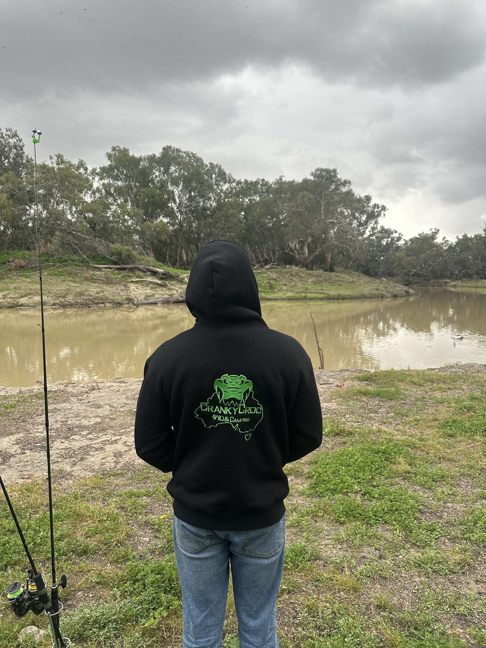 Person wearing a black hoodie with a green logo, standing by a river on a cloudy day.