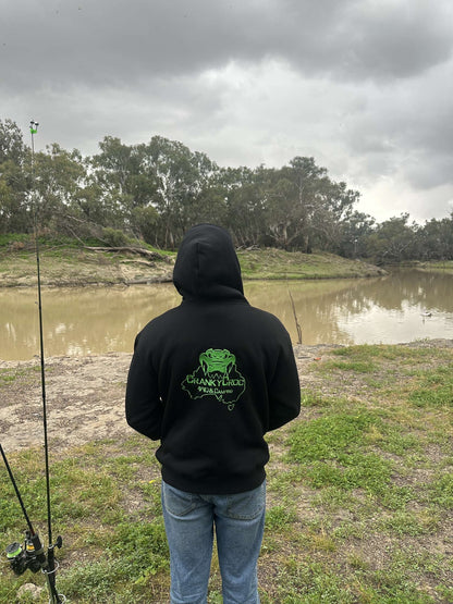 Person wearing a black hoodie with a green logo, standing by a river on a cloudy day.