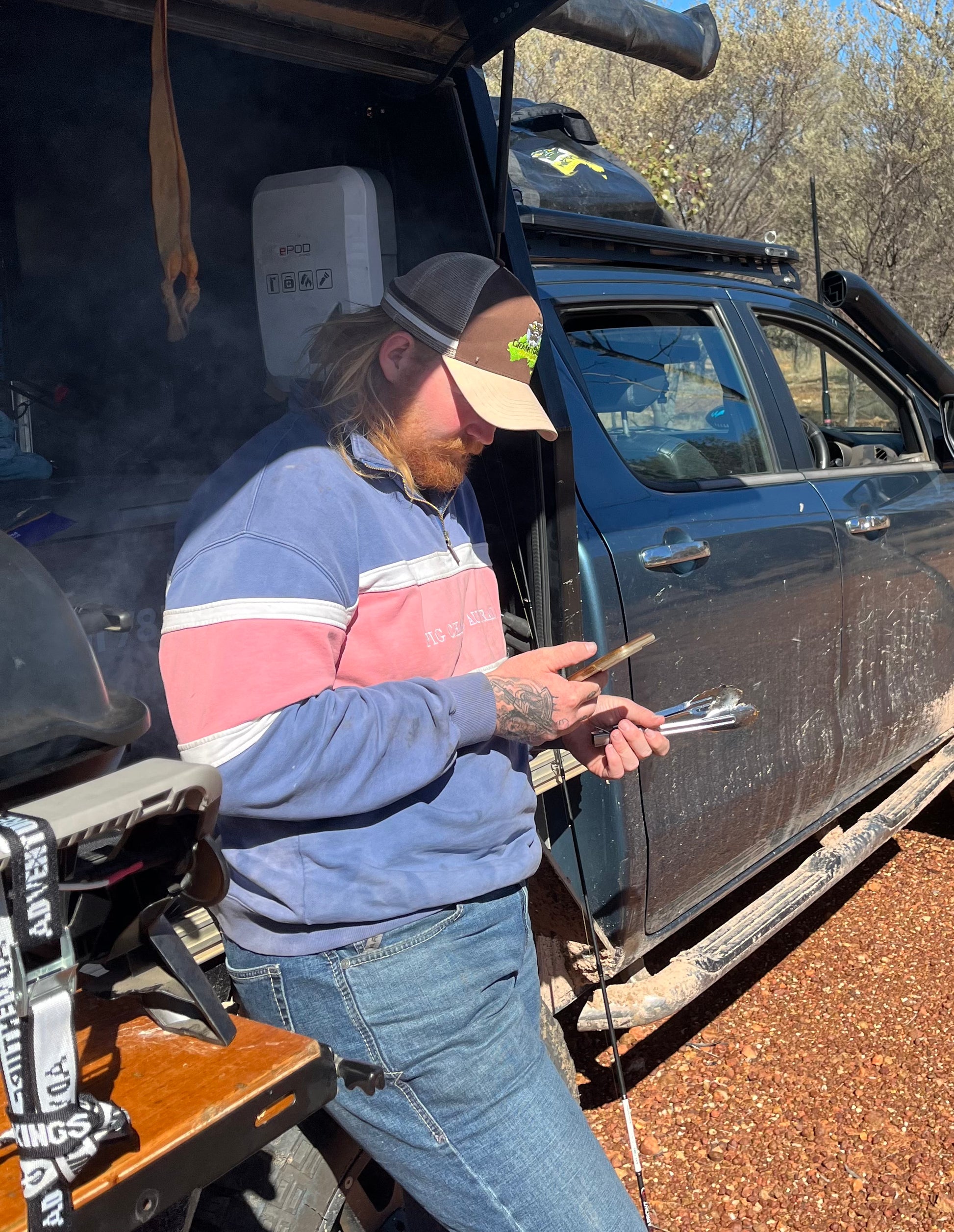 Man standing next to a vehicle holding a phone, the man is wearing a brown trucker hat. The trucker cap also has a brown and white stripe next on it. The image is set in outback Australia.