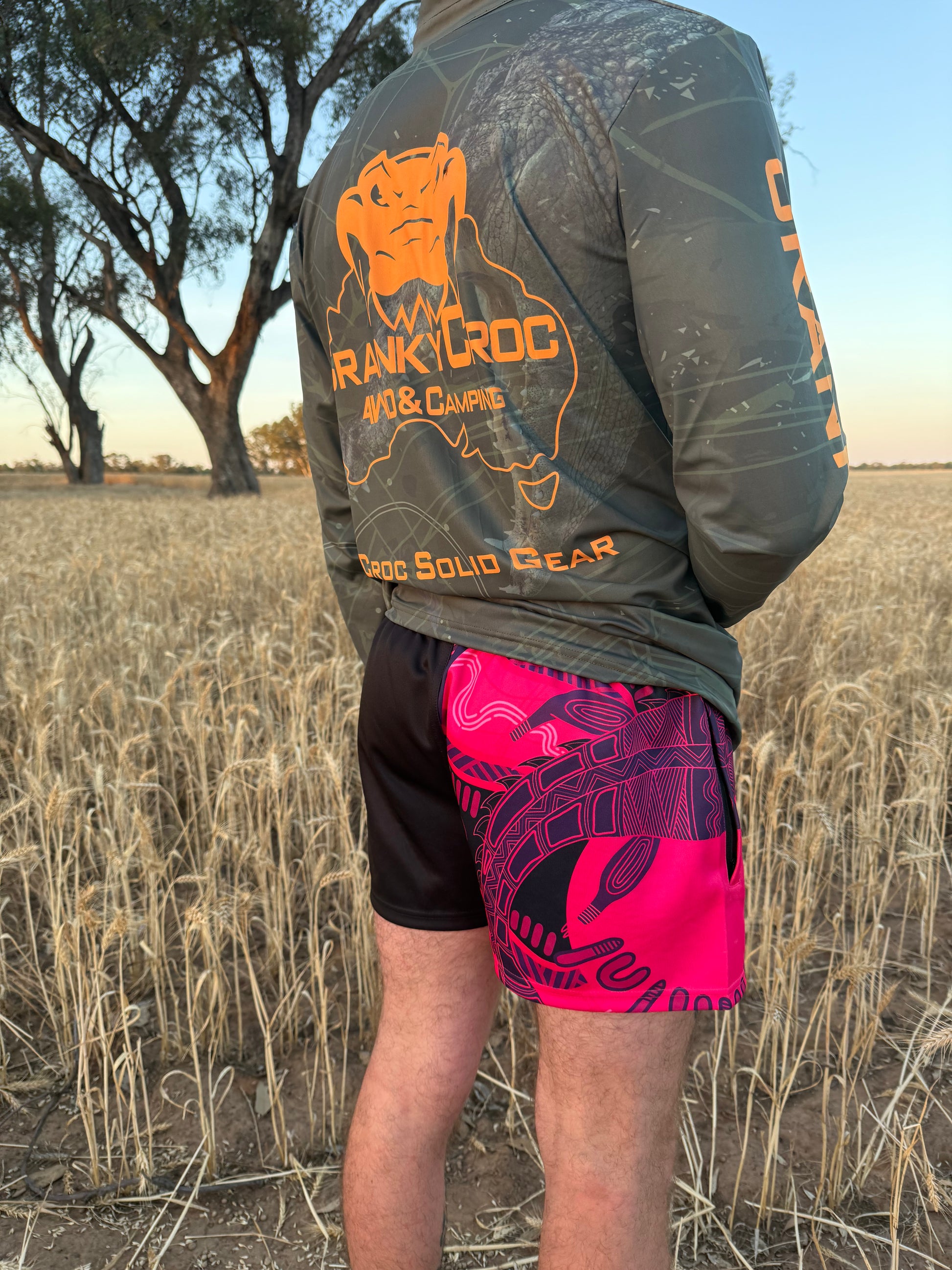Front view of person wearing Black and pink Indigenous Footy Shorts with zip pockets with a Cranky Croc logo and crocodile indigenous art design. Photo is set in a field